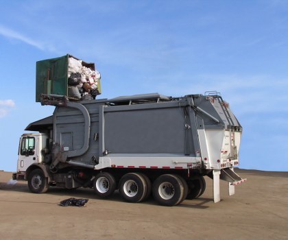 A large commercial rubbish collection truck is positioned on a paved area, with its rear loading compartment open to reveal a green container filled with mixed waste materials, including plastic bags and cardboard. The truck’s main body is grey with a metallic finish, contrasting with the white cab at the front. The vehicle has eight wheels on the visible side, and hydraulic lifting mechanisms are visible at the back, suggesting it is designed for waste collection. The scene is set outdoors against a clear blue sky with no other objects or structures nearby, indicating a disposal or collection site that could be typical around industrial or business areas in Canary Wharf or similar financial districts. The surface appears to be a flat, paved lot or driveway, with minimal environmental elements in view. The presence of waste materials and professional waste collection equipment aligns with services provided by Business Waste Removal Canary Wharf focused on commercial rubbish disposal in the local area.