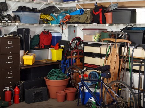 The image depicts a cluttered garage or storage area filled with various household items and tools. In the foreground, there are several empty terracotta plant pots of different sizes stacked and scattered on the floor. A blue bicycle with a black saddle and handlebars is leaning against a wooden storage rack on the right side of the image. Behind the bicycle, a black toolbox, a small black speaker, and a green garden hose are visible. On the left side, a black filing cabinet with four drawers sits next to a yellow plastic container and a red fire extinguisher positioned on the floor. The middle section features a metal shelving unit densely packed with boxes, plastic containers, and miscellaneous household items, including a red backpack, a grey tool bag, and a folded camping chair. The upper shelves hold additional boxes, ropes, and bags. The background includes more storage boxes and equipment, indicating an organized but crowded storage space typical of a family garage used for storing garden tools, outdoor equipment, and household goods associated with rubbish removal and general storage in the Canary Wharf area of London.